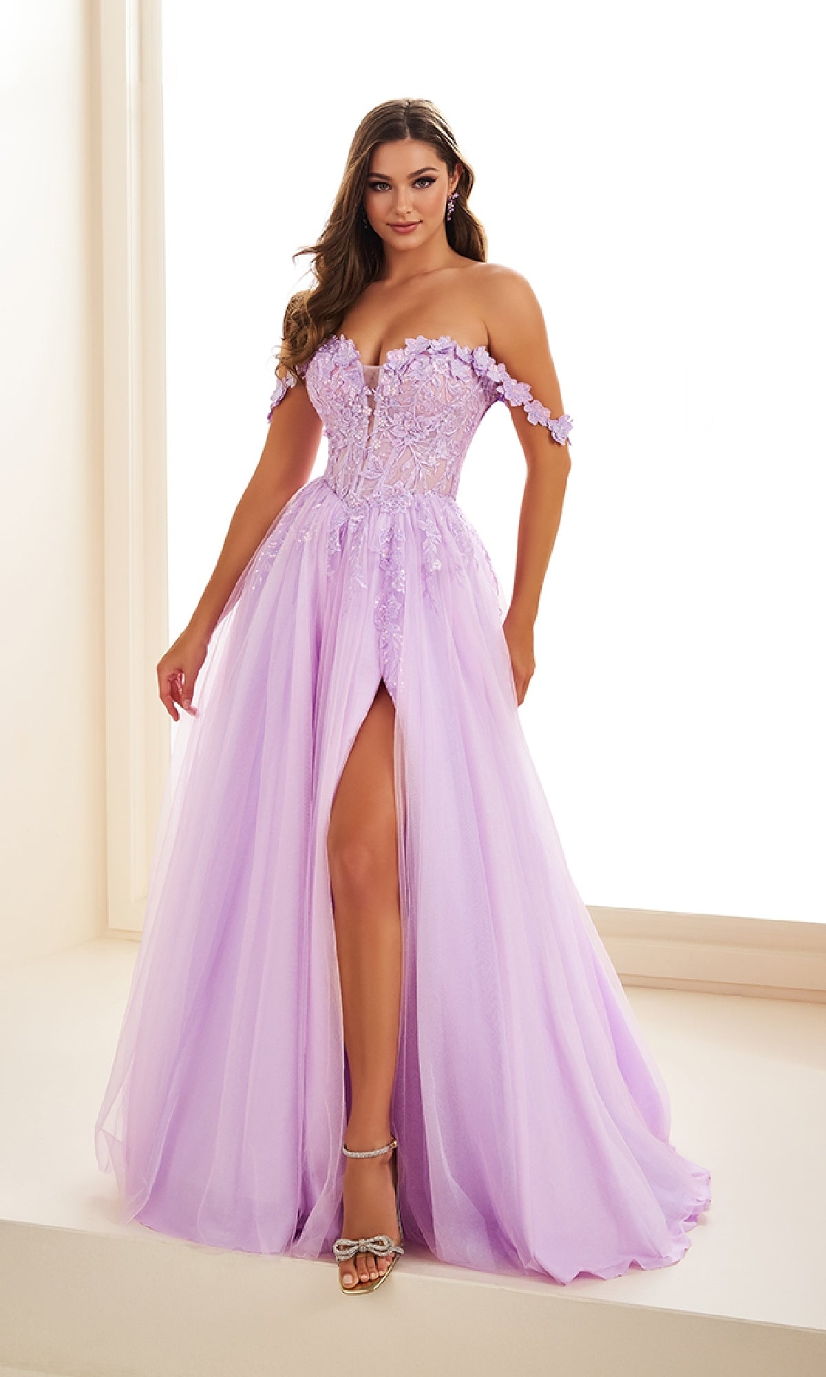 A woman stands in a bright room wearing the Ellie Wilde EW36214 long prom dress, featuring lavender tulle, an off-the-shoulder neckline, floral lace applique corset bodice, a-line silhouette, and a high slit.